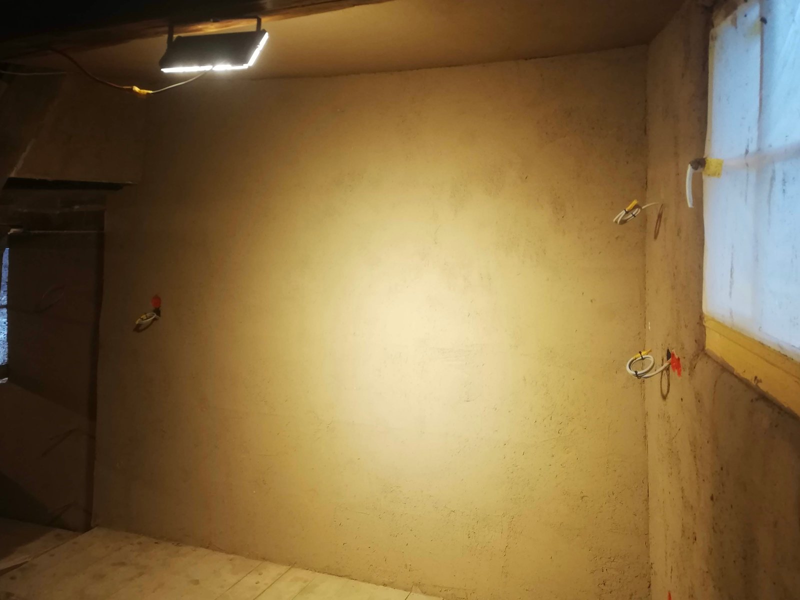 Lehmputz-Unterputz frisch aufgetragen, Baustrahler beleuchtet Wandfläche im Kellerraum